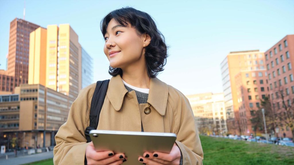 A young woman in a beige coat stands outdoors holding a tablet, with a backpack over her shoulder. She looks off to the side, smiling, in front of a cityscape with modern buildings and green grass.